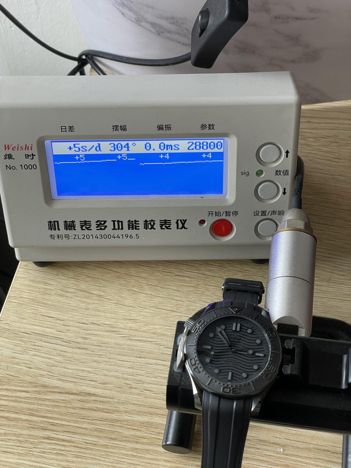 Single timegrapher screen beside Black Black Seamaster replica showing plus 5 seconds per day 304 degrees zero milliseconds and 28800